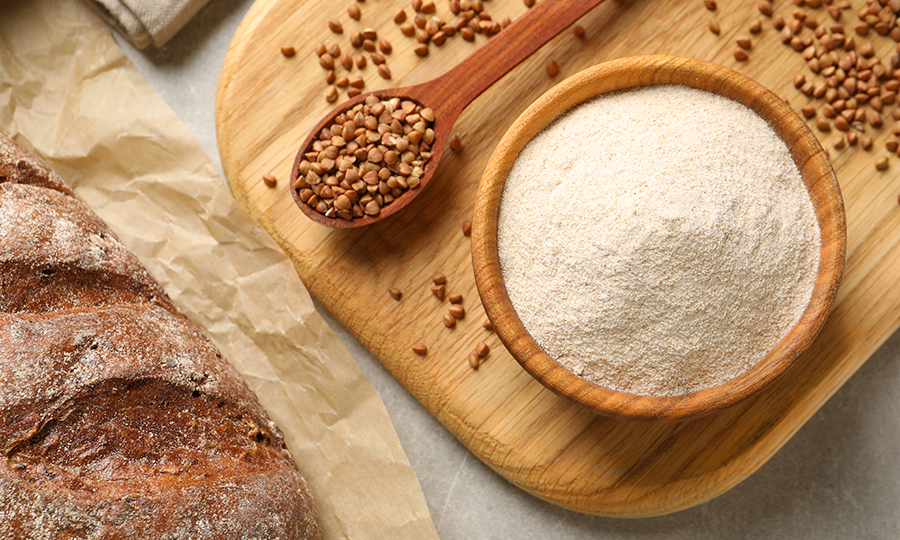 A wooden bowl of buckwheat flour with a spoonful of buckwheat seeds laid on a wooden table