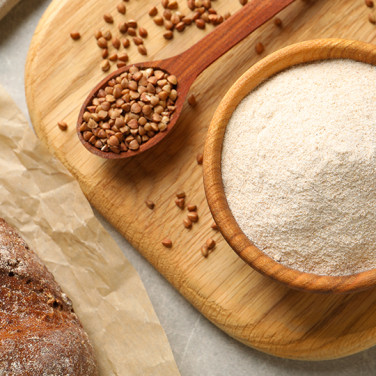 A wooden bowl of buckwheat flour with a spoonful of buckwheat seeds laid on a wooden table