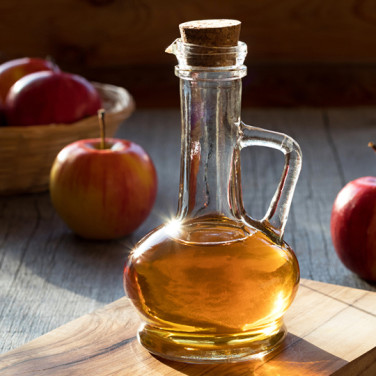 a small pitcher of apple cider vinegar hit with direct sunlight on wooden cutting board and apples in the back