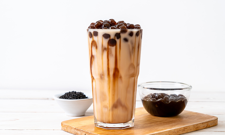 A glass of brown sugar bubble tea on a small wooden platter next to a small bowl of tapioca pearls