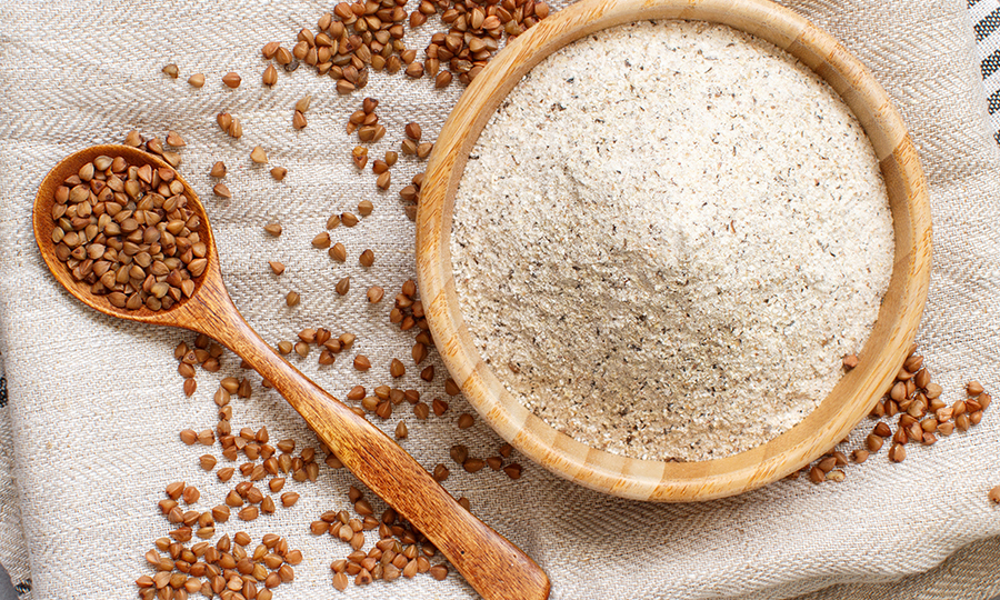 A big wooden bowl of buckwheat flour next to a small wooden spoon filled with buckwheat seeds