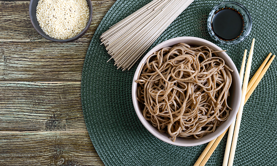 A bowl of soba buckwheat noodles next to some sauce, uncooked buckwheat noodles, and chopsticks