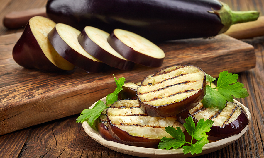 Grilled slices of eggplant plated on a recyclable dish in front of a sliced raw eggplant and a uncut eggplant