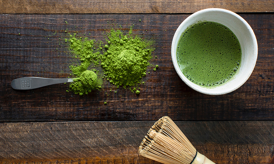 From left to right, an overflowing metal spoonful of matcha powder next to some freshly brewed matcha tea and a matcha brush
