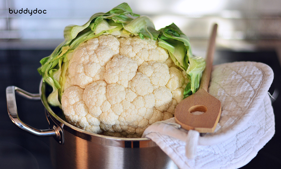 cauliflower head in brass pot with water next to wooden spatula and kitchen rag