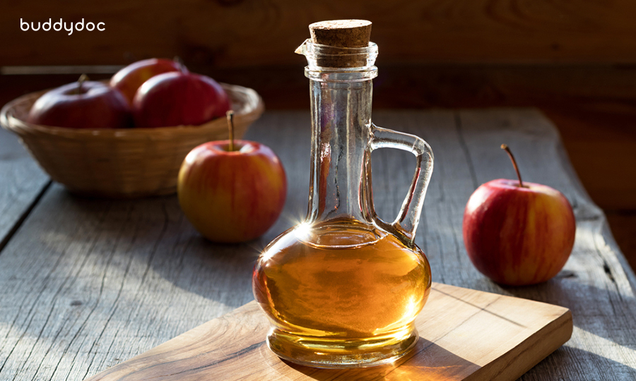 a small pitcher of apple cider vinegar hit with direct sunlight on wooden cutting board and apples in the back